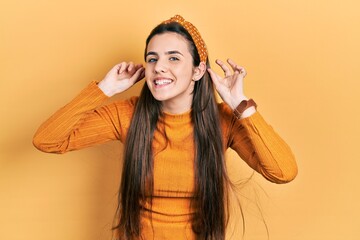 Young brunette teenager wearing casual yellow sweater smiling pulling ears with fingers, funny...