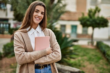 Young hispanic student girl smiling happy holding book at the city.