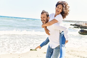Middle age hispanic man smiling happy holding woman on his back at the beach. © Krakenimages.com