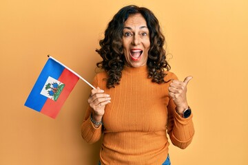 Middle age hispanic woman holding haiti flag pointing thumb up to the side smiling happy with open mouth