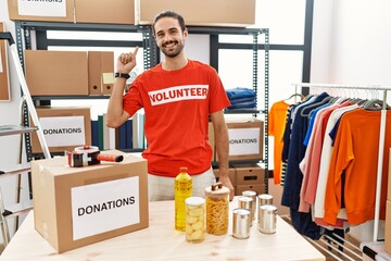 Young hispanic man wearing volunteer t shirt at donations stand cheerful with a smile on face...