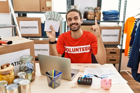 Young hispanic man holding money at donations stand smiling with an idea or question pointing finger with happy face, number one