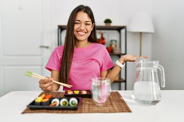 Young brunette woman eating sushi using chopsticks smiling cheerful presenting and pointing with palm of hand looking at the camera.