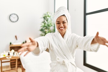 Young brunette woman wearing towel and bathrobe standing at beauty center looking at the camera smiling with open arms for hug. cheerful expression embracing happiness.