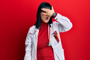 Young hispanic woman wearing doctor uniform and stethoscope peeking in shock covering face and eyes with hand, looking through fingers with embarrassed expression.