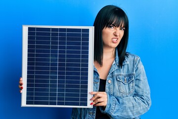 Young hispanic woman holding photovoltaic solar panel clueless and confused expression. doubt concept.