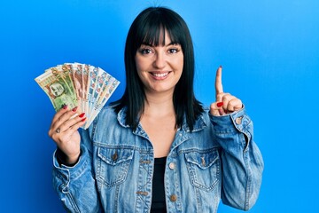Young hispanic woman holding peruvian sol banknotes smiling with an idea or question pointing finger with happy face, number one