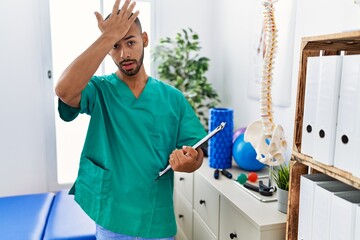 Fototapeta premium African american physiotherapist man working at pain recovery clinic surprised with hand on head for mistake, remember error. forgot, bad memory concept.