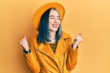 Young modern girl wearing yellow hat and leather jacket very happy and excited doing winner gesture with arms raised, smiling and screaming for success. celebration concept.