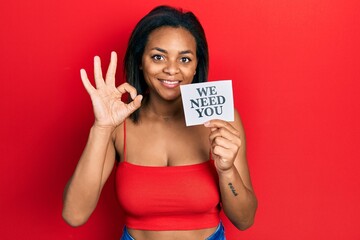 Young african american girl holding we need you paper doing ok sign with fingers, smiling friendly gesturing excellent symbol