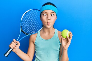 Beautiful brunette little girl playing tennis holding racket and ball making fish face with mouth...