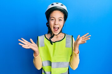 Beautiful brunette little girl wearing bike helmet and reflective vest celebrating crazy and amazed for success with arms raised and open eyes screaming excited. winner concept