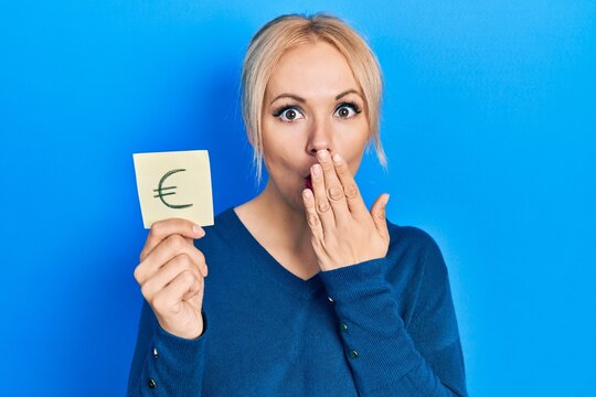 Young blonde woman holding euro symbol reminder covering mouth with hand, shocked and afraid for mistake. surprised expression