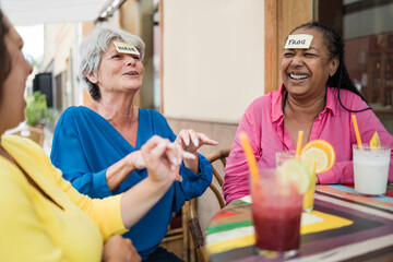 Multiracial senior friends having fun playing together guess who forehead game at bar restaurant -...