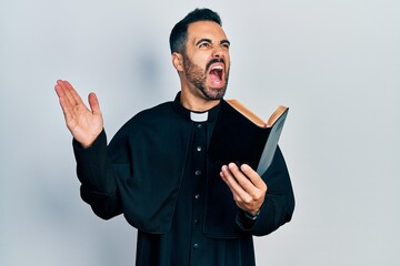 Handsome hispanic priest man with beard holding bible and christian cross angry and mad screaming...
