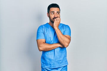 Handsome hispanic man with beard wearing blue male nurse uniform looking stressed and nervous with hands on mouth biting nails. anxiety problem.