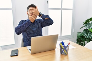 Young hispanic man with beard working at the office with laptop covering eyes and mouth with hands, surprised and shocked. hiding emotion