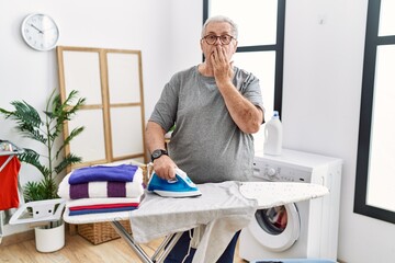 Senior caucasian man ironing clothes at home covering mouth with hand, shocked and afraid for mistake. surprised expression