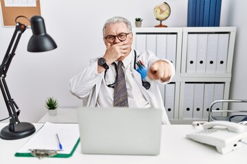 Senior caucasian man wearing doctor uniform and stethoscope at the clinic laughing at you, pointing finger to the camera with hand over mouth, shame expression