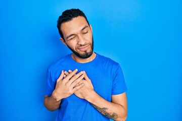 Hispanic man with beard wearing casual blue t shirt smiling with hands on chest with closed eyes and grateful gesture on face. health concept.