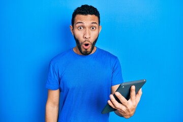 Hispanic man with beard using touchpad device scared and amazed with open mouth for surprise, disbelief face