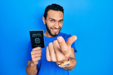 Hispanic man with beard holding italy passport beckoning come here gesture with hand inviting welcoming happy and smiling