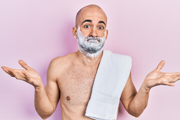 Young bald man shirtless shaving beard with foam clueless and confused expression with arms and...