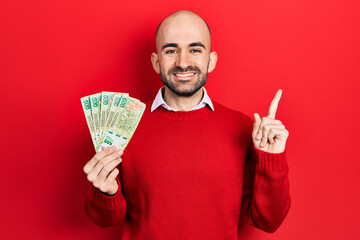 Young bald man holding argentine pesos banknotes smiling happy pointing with hand and finger to the...