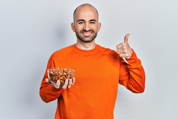 Young bald man holding peanuts smiling happy and positive, thumb up doing excellent and approval sign