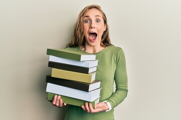 Young blonde woman holding a pile of books celebrating crazy and amazed for success with open eyes screaming excited.