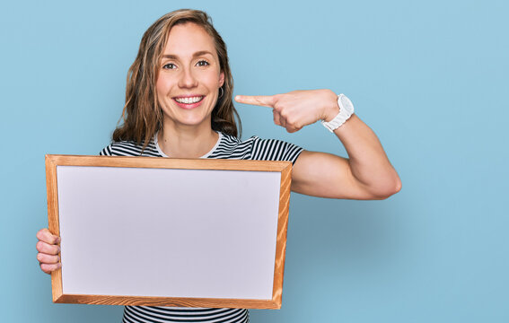 Young blonde woman holding empty white board pointing finger to one self smiling happy and proud