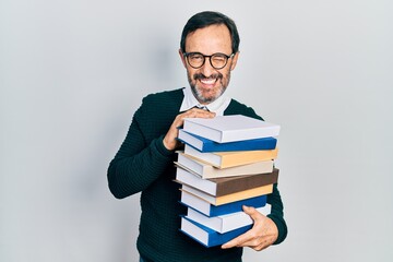 Middle age hispanic man holding a pile of books winking looking at the camera with sexy expression, cheerful and happy face.