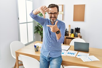 Middle age hispanic man with beard wearing business clothes at the office smiling making frame with...