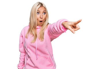 Young blonde woman wearing casual sweatshirt pointing with finger surprised ahead, open mouth amazed expression, something on the front