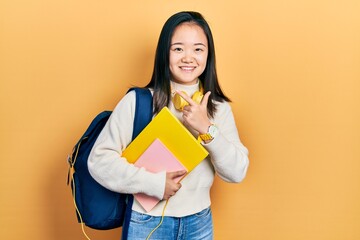 Young chinese girl holding student backpack and books cheerful with a smile on face pointing with hand and finger up to the side with happy and natural expression