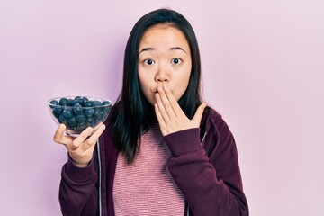 Young chinese girl holding blueberries covering mouth with hand, shocked and afraid for mistake....