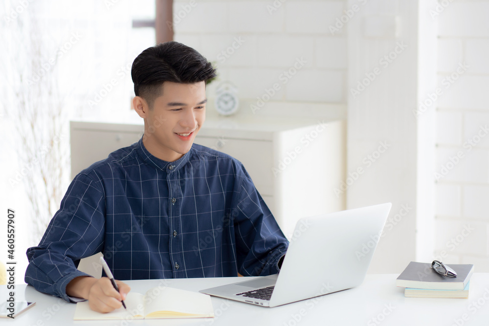 Sticker Young asian business man writing on notebook for planning working and using laptop computer on desk at home, notes about finance, male study and learning, business and communication concept.