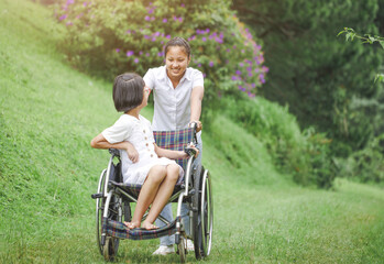 Disabled child sitting in wheelchair and care helper walking on meadow park in sunny day. International Disability Day.