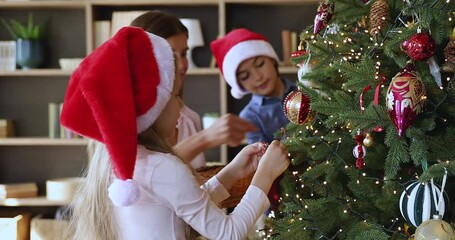 Family Christmas tradition. Happy young mom having fun with son daughter kids at Xmas Eve trim New Year tree prepare to celebrate holiday. Smiling nanny assist children to decorate fir tree with toys