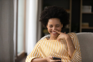 Laughing millennial generation mixed race african ethnicity woman holding smartphone in hands, watching entertaining video or photo content, reading message with good news, having fun at home.
