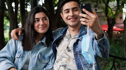 A young Hispanic couple in a park taking a cell phone picture, wearing masks. Health care