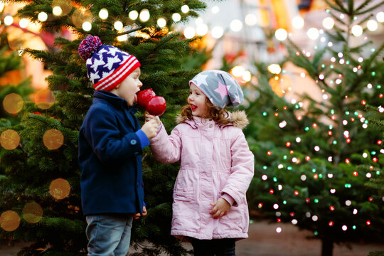 Two little smiling kids, preschool boy and girl eat sweet sugared apple on German Christmas market. Happy siblings children with lights on background and xmas trees. Family funny brother and sister.