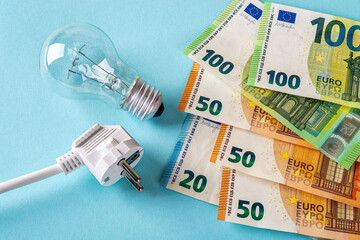 White electric plug, light bulb and euro money banknotes over blue background. Increasing of electricity cost expensive energy bill and rise in electricity prices concepts. 