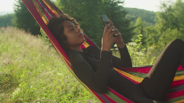 Side View Of Peaceful Happy Pretty African American Woman Lying And Swaying In Hammock, Networking And Browsing Social Media Content Online On Mobile Phone While Enjoying Summer Vacations In Mountains