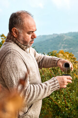 Bearded man in woolen sweater pouring hot coffee or tea from thermos bottle in a cup in autumn garden with colorful fall nature in background. Fall mood. People in autumn.