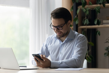Smiling businessman in glasses using smartphone at workplace, successful confident man entrepreneur freelancer sitting at home office desk, typing writing message in social network, browsing apps