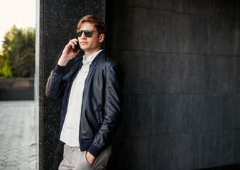 Portrait of a handsome young businessman in black sunglasses and leather jacket, talking on a cell phone, outside.