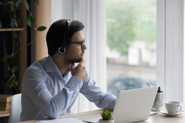 Thoughtful man in glasses and headphones looking in distance, sitting at work desk with laptop,...