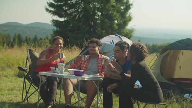 Cheerful happy diverse multicultural backpackers sitting at camping table, toasting with travel mugs, celebrating successful mountain hiking at campsite in morning while enjoying recreation on trek.