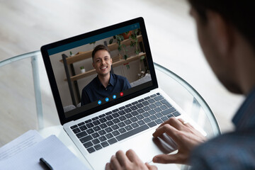 Fototapeta na wymiar View over businessman freelancer shoulder chatting online by video call, smiling colleague or mentor on laptop screen speaking, young man engaged in internet meeting or interview by webcam
